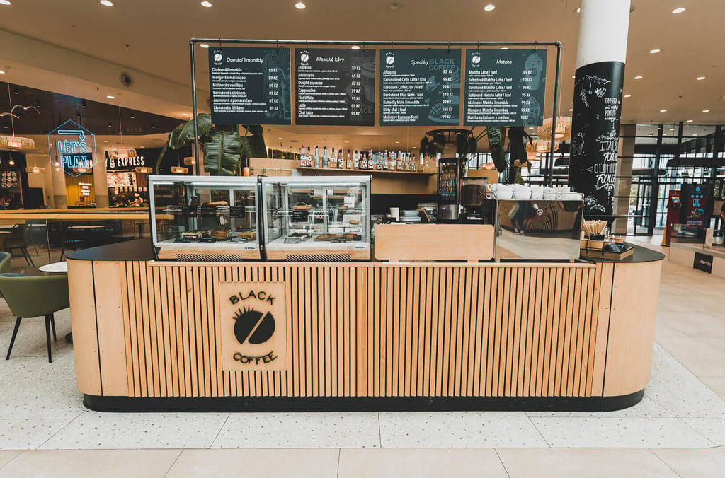 Dřevěný bar Black Coffee s prosklenou vitrínou na dezerty a nasvícenou menu tabulí v interiéru pasáže